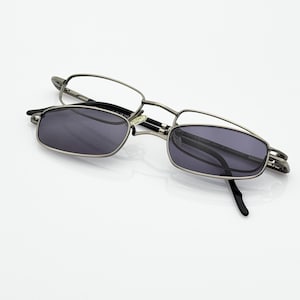 Op de afbeelding: Een bril met zilveren montuur en een donkergrijze zonnebril met clip-on. De bril heeft een rechthoekige vorm en staat tegen een witte achtergrond. De glazen zijn licht getint.