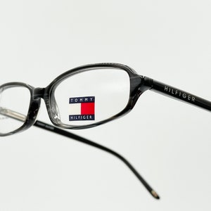 May include: Black and gray rectangular eyeglasses with a Tommy Hilfiger logo on the right lens. The brand name "HILFIGER" is printed on the right arm of the glasses.