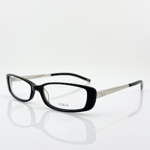 Puede incluir: Gafas rectangulares negras con varillas plateadas y la marca "FURLA" impresa en la montura. Las gafas están sobre un fondo blanco, con un reflejo visible debajo.