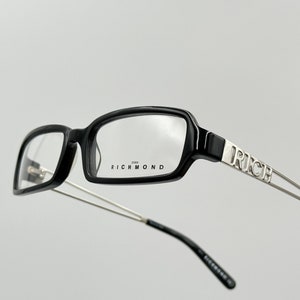 Puede incluir: Gafas rectangulares negras con patillas plateadas. Las patillas están grabadas con la palabra "RICH" y el nombre de la marca "JOHN RICHMOND" está impreso en la montura.