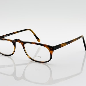 May include: Eyeglasses with a tortoiseshell pattern. The frames are brown and black, with clear lenses. The brand name "Compa" is visible on the frame. The glasses have a classic, slightly rounded shape.