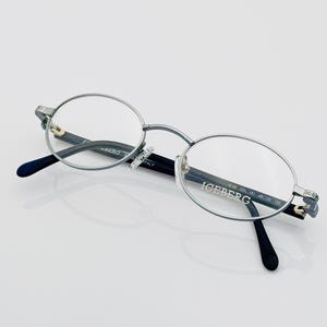 Puede incluir: Gafas redondas plateadas con patillas negras y la palabra "ICEBERG" impresa en la patilla derecha.