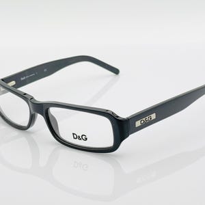 Puede incluir: Gafas rectangulares negras con lentes transparentes. Las monturas presentan el logotipo "D&G" en las patillas y en el interior de la lente izquierda. Las gafas están sobre un fondo blanco, con un reflejo visible debajo.