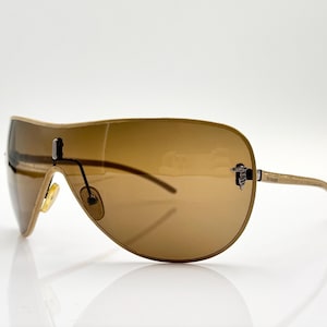 Pode incluir: Óculos de sol aviador marrons com armação dourada e lentes marrons. Os óculos têm um design único com uma armação curva e uma fina barra de metal que conecta as lentes.