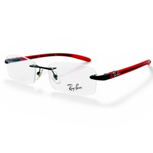 May include: Rimless eyeglasses with clear lenses and red temples. The Ray-Ban logo is visible on the lens. The temples have a black stripe and the Ray-Ban logo. The glasses have a modern, sporty design.
