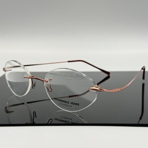 May include: A pair of rimless eyeglasses with a rose gold frame and clear lenses. The brand name "FACONNABLE JEANS EYEWEAR" is printed on the right lens.