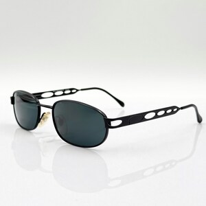 May include: Black oval sunglasses with dark lenses. The frames are black metal with a decorative oval design on the temples. The sunglasses are set against a white background, with a reflection visible below.