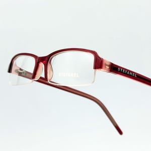 Puede incluir: Gafas STEFANEL rojas con un diseño semi-sin montura. Las monturas son de color rojo intenso, con el nombre de la marca impreso en la varilla. Las lentes son transparentes y el estilo general es moderno y elegante.