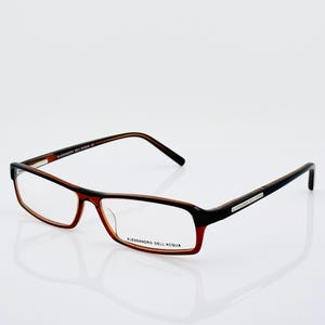 May include: Alessandro Dell'Acqua eyeglasses featuring a rectangular frame with a two-tone design. The upper portion of the frame is black, transitioning into a warm amber-brown hue at the bottom. The brand name is printed on the lens and temple.