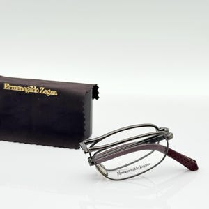 Può includere: Occhiali pieghevoli con montatura in metallo scuro e dettagli bordeaux. Gli occhiali sono accanto a un astuccio marrone scuro con la scritta dorata "Ermenegildo Zegna". Gli occhiali sono parzialmente piegati.