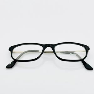 May include: Black-framed eyeglasses with a rectangular shape and clear lenses. The glasses have a thin, silver-colored bridge and temples. The brand name "Ganna" is visible on one lens. The glasses are set against a white background.