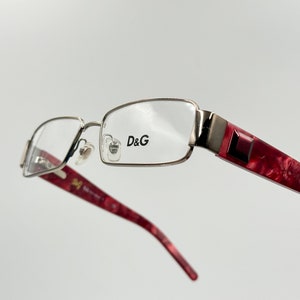 Puede incluir: Gafas de metal plateado con lentes transparentes y patillas de plástico rojo con efecto marmoleado. El nombre de la marca "D&G" está impreso en la montura.
