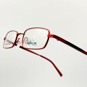 Puede incluir: Gafas de metal con montura roja y lentes transparentes. El nombre de la marca "Kipling Eyewear" es visible en la lente derecha.