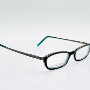 Op de afbeelding: Zwarte en teal rechthoekige bril met dunne, donkergrijze metalen pootjes. De monturen hebben een zwarte buitenrand met teal accenten. De merknaam "MARIUS MOREL" staat op het montuur.