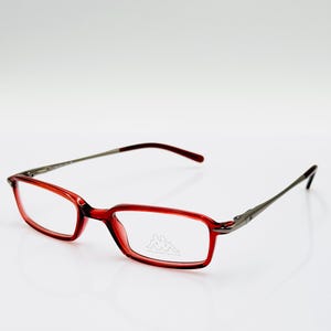 Puede incluir: Gafas rectangulares rojas con varillas plateadas. Las monturas son de color rojo translúcido y las lentes son transparentes. El logotipo de Kappa es visible en la lente derecha.