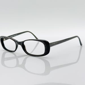 Puede incluir: Gafas negras con montura rectangular y lentes transparentes. Las patillas son de color gris oscuro con el nombre de la marca "ICEBERG". Las gafas están sobre un fondo blanco, proyectando una sombra.