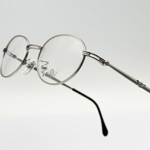 May include: Silver-toned round eyeglasses with thin metal frames and clear lenses. The arms of the glasses have a decorative design.