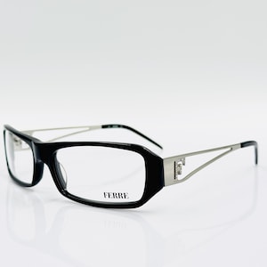 Puede incluir: Gafas rectangulares negras con patillas de metal plateado y un detalle de metal plateado en la parte delantera de la montura. El nombre de la marca "FERRE" está impreso en la lente derecha.