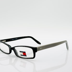 May include: Rectangular eyeglasses with black frames and gray temples. The Tommy Hilfiger logo is visible on the left lens. The glasses are set against a white background.