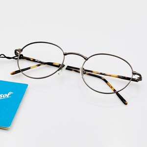 May include: A pair of round, silver-toned spectacles with tortoiseshell temple arms. The glasses are on a white surface with a blue tag that says "Persol" in white letters.