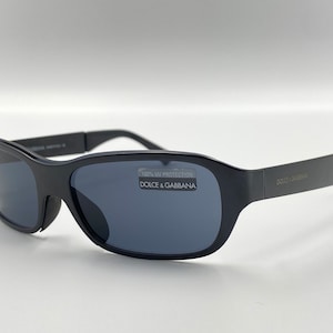 May include: Black sunglasses with a dark gray lens and a black frame. The sunglasses have a label on the right lens that says "100% UV Protection Dolce & Gabbana".