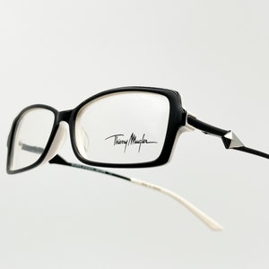 May include: Black and white rectangular eyeglasses with a silver accent on the temple. The brand name "Thierry Mugler" is printed on the right lens.