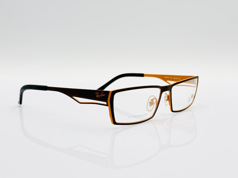 K&ouml;nnte beinhalten: Schwarze Brille mit goldfarbenem Rahmen und Titan-Gestell. Die Brille hat eine rechteckige Form und klare Gl&auml;ser.