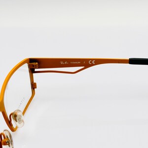 K&ouml;nnte beinhalten: Orangefarbene Metallgestellbrille mit schwarzen B&uuml;gelenden. Das Gestell tr&auml;gt den Schriftzug "Ray-Ban TITANIUM Z CE" an der Seite.