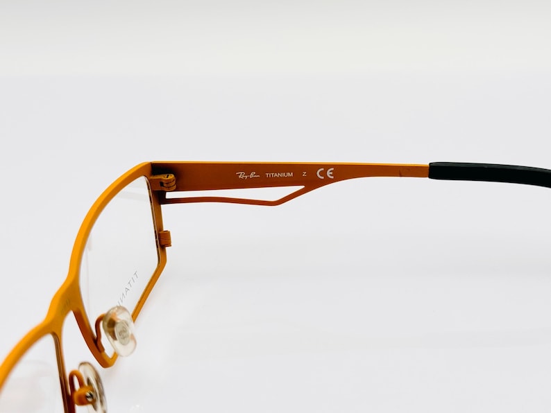 K&ouml;nnte beinhalten: Orangefarbene Metallgestellbrille mit schwarzen B&uuml;gelenden. Das Gestell tr&auml;gt den Schriftzug "Ray-Ban TITANIUM Z CE" an der Seite.