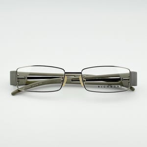 Puede incluir: Gafas de montura rectangular en gris y plata. Las gafas tienen un diseño minimalista con la marca "RICHMOND" visible en la patilla derecha. Las lentes son transparentes. La anchura de la montura es de aproximadamente 13,2 cm.
