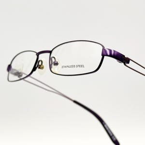 May include: Purple metal frame eyeglasses with clear lenses. The frame has the words "Stainless Steel" printed on the right lens.