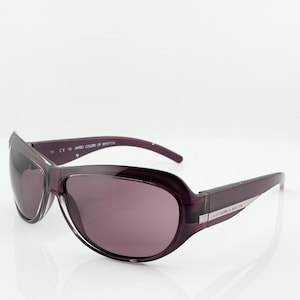 Puede incluir: Gafas de sol de color púrpura oscuro con montura curvada y lentes tintadas. Las gafas de sol tienen el texto "UNITED COLORS OF BENETTON" impreso en la patilla. Las lentes son de un tono púrpura a juego.
