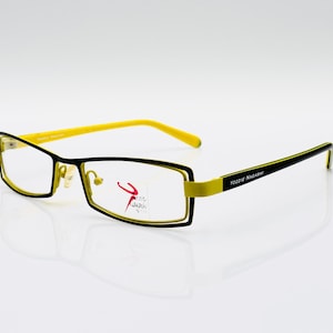 Puede incluir: Gafas rectangulares negras y amarillas. El nombre de la marca "TOZZIE NAGANO" está impreso en la patilla derecha.