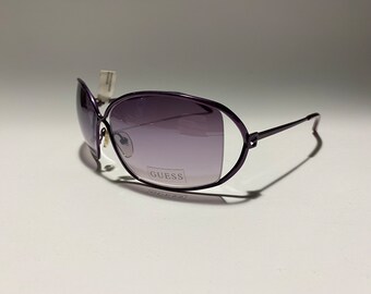 Puede incluir: Gafas de sol Guess moradas con lentes degradados. Las gafas tienen una forma ovalada redondeada y un fino marco de metal.
