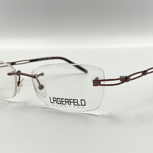 May include: Brown rimless eyeglasses with a metal frame and a double-bridge design. The brand name "LAGERFELD Lunettes" is printed on the right lens.