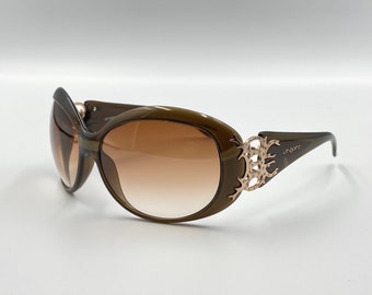 Puede incluir: Gafas de sol ovaladas con lentes degradadas marrones y montura verde oliva oscuro. Las patillas presentan un adorno decorativo de color dorado. El nombre de la marca "ungaro" está impreso en la patilla.