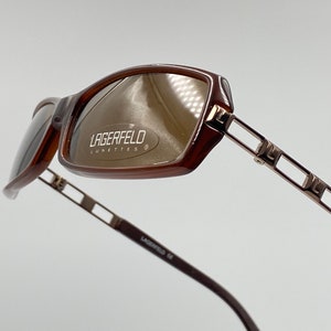 May include: Brown and gold-toned sunglasses with a rectangular frame and a silver metal temple with the text "LAGERFELD lunettes" on the lens.