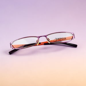 Può includere: Occhiali viola con dettagli arancioni e il marchio "MORGAN" visibile sulla lente. Gli occhiali hanno una montatura rettangolare e sono su uno sfondo sfumato viola e arancione.