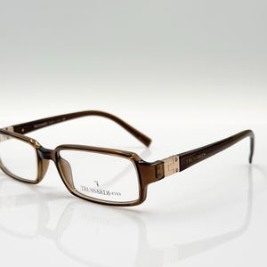 Puede incluir: Gafas de pasta marrón con lentes transparentes. El nombre de la marca "TRUSSARDI EYES" está impreso en la patilla derecha.