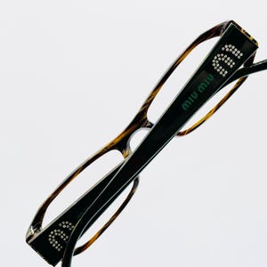 May include: Eyeglasses with a dark brown and tortoise shell frame. The temples are black with the brand name "MIU MIU" in white and the letters "MIU" in rhinestones. The glasses are set against a white background.