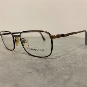 Puede incluir: Gafas de metal de color marrón con lentes transparentes. El nombre de la marca "EMPORIO ARMANI" está impreso en la lente derecha.