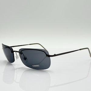 Peut inclure: Lunettes de soleil sans monture avec verres gris foncé et monture fine en métal noir. Les verres rectangulaires sont légèrement incurvés et les branches sont droites. Les lunettes sont posées sur un fond blanc, avec un reflet visible en dessous.