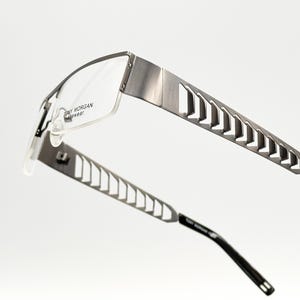 Op de afbeelding: Zilverkleurige metalen bril met een rechthoekig montuur en een uitgesneden ontwerp op de pootjes. De merknaam "TONY MORGAN EYEWEAR" is op het montuur gedrukt.