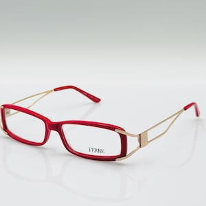 Puede incluir: Gafas rojas y doradas con montura rectangular. Las gafas tienen una montura roja llamativa con varillas y patillas doradas. El nombre de la marca "FERRE" está impreso en la lente. Estas gafas son un accesorio elegante.