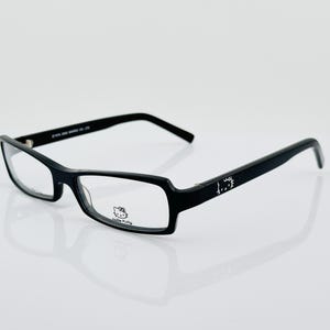 May include: Black rectangular eyeglasses with clear lenses. The frames feature a Hello Kitty logo on the lower left lens and the Hello Kitty logo on the temple. The glasses are set against a white background.