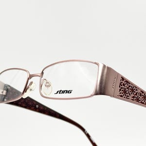 Può includere: Occhiali da vista in oro rosa con un design floreale sulle aste e la parola "STING" stampata sulla montatura.