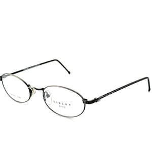 Puede incluir: Gafas ovaladas con montura gris oscuro y varillas negras. La marca "SISLEY" es visible en la montura. Las lentes son transparentes y las gafas están sobre un fondo blanco.