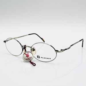 May include: Silver-toned eyeglasses with oval lenses and black temples. The frames feature the Playboy logo on the lens and a charm with the Playboy bunny logo. The glasses are set against a white background.