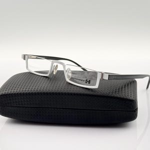 Può includere: Occhiali rettangolari argentati e neri con una custodia nera. Gli occhiali hanno una montatura argentata e lenti trasparenti. La custodia è realizzata in tessuto nero con un motivo testurizzato.