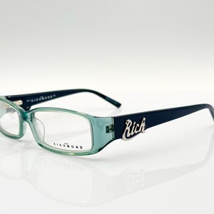 Puede incluir: Gafas verdes y negras con un logotipo "Rich" plateado en la patilla derecha. El nombre de la marca "Richmond" está impreso en la patilla izquierda.